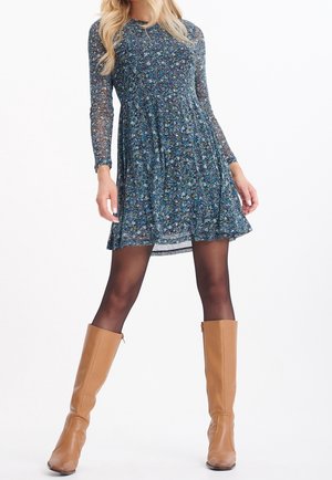 Donna che indossa un vestito lungo a maniche lunghe con motivi floreali blu, collant neri trasparenti e stivali beige al ginocchio, in piedi con le gambe divaricate su sfondo bianco.