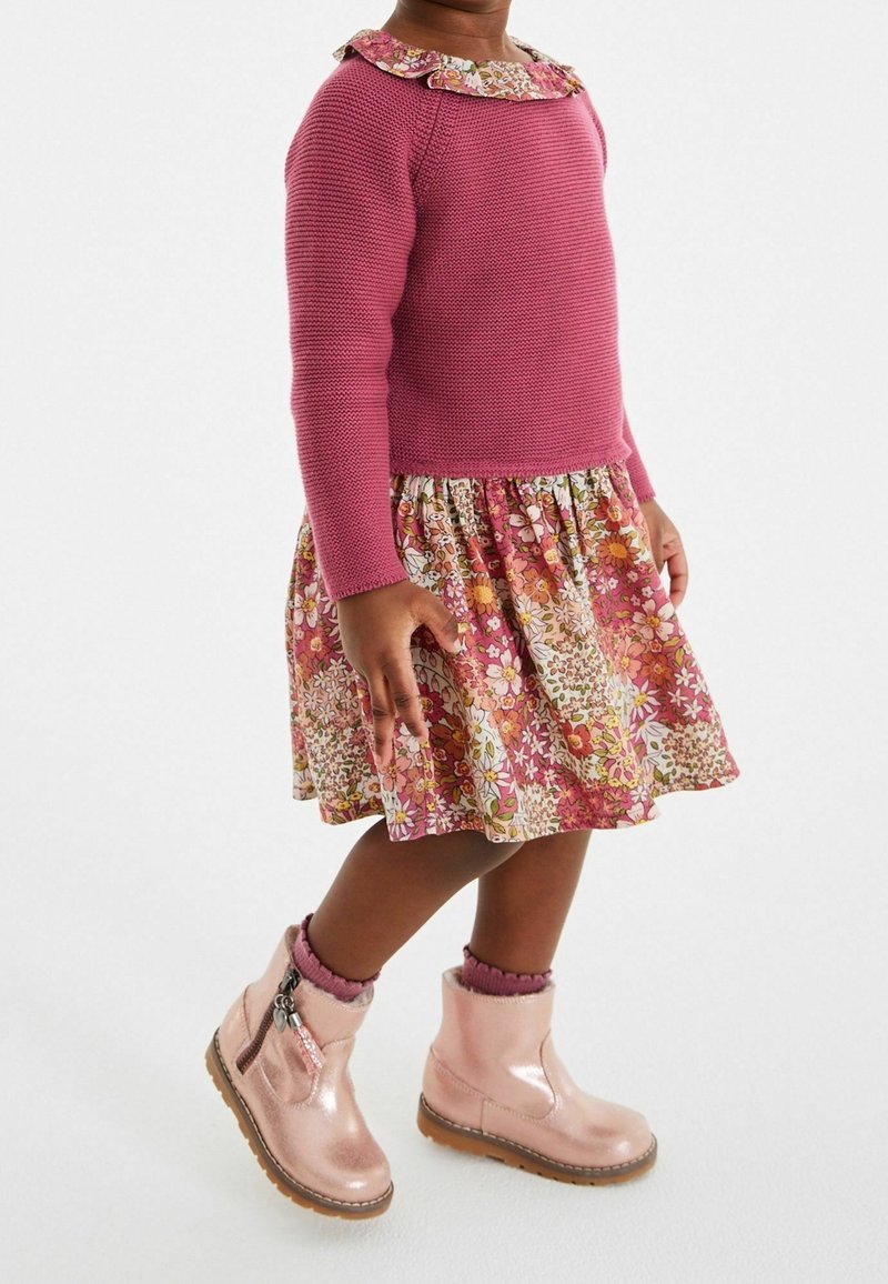 Enfant portant un pull en tricot rose, une robe fleurie avec un col à volants, des chaussettes roses et des bottes roses claires brillantes, debout sur un fond blanc.