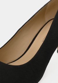 Escarpin à talon haut en daim noir à finition lisse, présentant un bout arrondi et un talon fin. Intérieur beige clair avec des détails subtils.