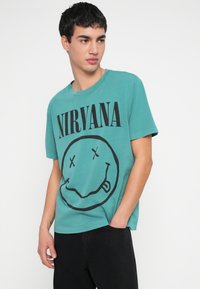 Τυρκουάζ βαμβακερό t-shirt με μεγάλο μαύρο γραφικό χαμογελαστού προσώπου και το κείμενο "NIRVANA" σε έντονα μαύρα γράμματα. Λαιμόκοψη crew neck και κοντά μανίκια.