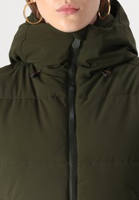Olijf groene gewatteerde jas met hoge kraag, ritssluiting en verstelbare trekkoorden. Heeft een textiele stof en metalen accenten.