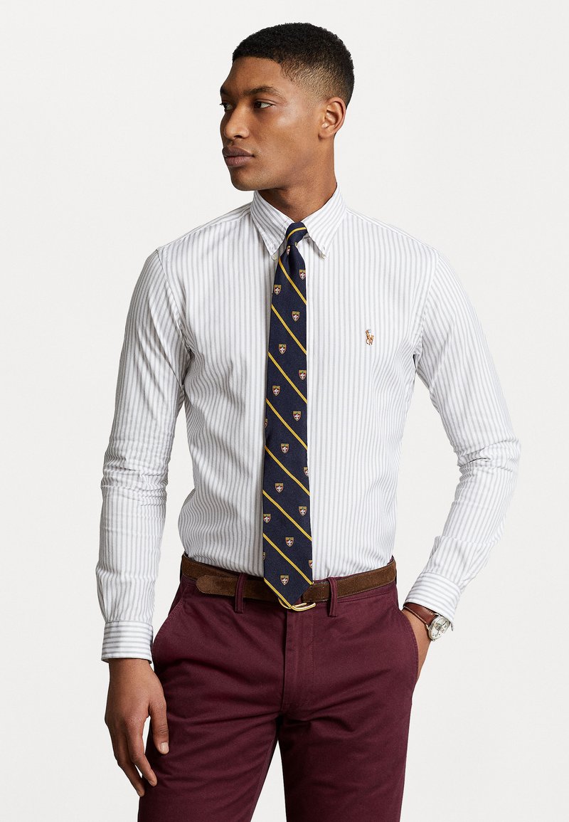 Cămașă albă și albastră cu dungi, cu guler, asociată cu o cravată bleumarin cu embleme patterned. Pantaloni bordeaux și o curea maro.