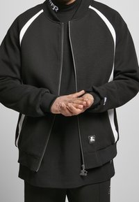 Chaqueta bomber negra con rayas blancas, cierre de cremallera, puños acanalados y etiqueta con logo. Fabricada con tela suave y de corte holgado.