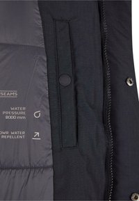 Musta takki, jonka sisäosassa on tilkkukangasta, sivutasku, vedenpainelappu ja oikealla puolella vetoketju turvallista kiinnioloa varten.