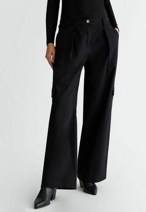 Pantaloni neri a gamba larga realizzati in tessuto morbido, con dettagli a pieghe e tasche laterali. Il modello indossa stivali neri con tacco.