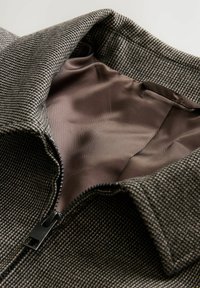 Chaqueta gris texturizada con cremallera negra y forro marrón. El cuello está doblado, revelando la suave tela interior.
