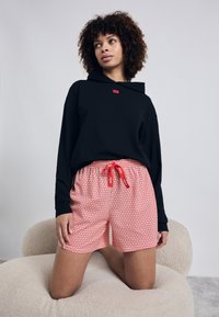 Zwarte sweatshirt met capuchon en rood logo, gecombineerd met rode shorts met patroon, die een elastische tailleband en rode trekkoord hebben.