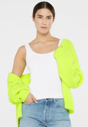 Neon-grüner, oversized Cardigan mit gerippten Bündchen, getragen über einem weißen Tanktop. Der Cardigan hat eine weiche Textur und einen lockeren Schnitt.