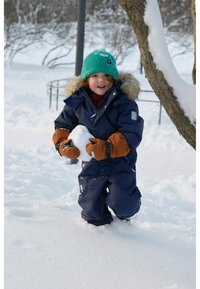 Marine sneeuwpak met bont afgewerkte capuchon, bruine wanten en groene gebreide muts. Kind houdt een sneeuwbal vast en staat in diepe sneeuw.