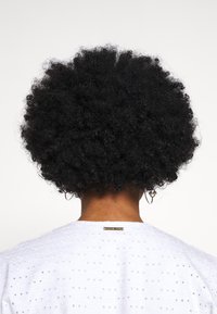 Capelli neri ricci a forma arrotondata, portati sciolti. Abbigliamento bianco con motivi floreali a filetto e un'etichetta di marca sul retro.