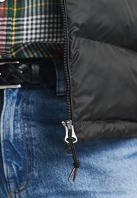 Svart puffjacka som är delvis zippad över blå jeans, svart flätad bälte, och en grön, röd och vit rutad skjorta.