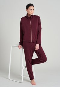 Burgundská souprava domácího oblečení s bundou na zip a vysokým límcem a odpovídajícími kalhotami, vyrobená z měkké tkaniny a s volným střihem.