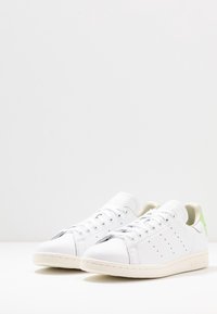 Ett par vita låga sneakers med perforerade sidor, vita snören, beige sulor och ljusgröna häftplåster på en vit yta.