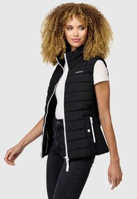 Gilet rembourré noir avec un col montant, fermeture éclair et accents blancs. Présente des poches latérales et une texture matelassée. Porté par-dessus une chemise blanche.