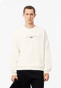 Felpa color crema con scollo rotondo, caratterizzata dalla scritta "LACOSTE" ricamata in nero e dal logo di coccodrillo verde. Maniche lunghe e polsini a costine.