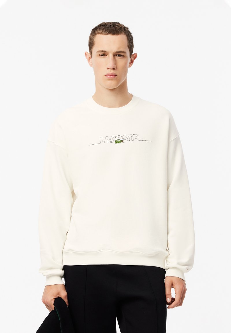 Felpa color crema con scollo rotondo, caratterizzata dalla scritta "LACOSTE" ricamata in nero e dal logo di coccodrillo verde. Maniche lunghe e polsini a costine.