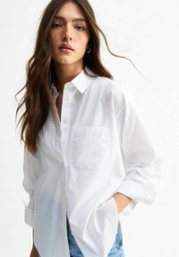 Chemise boutonnée en coton blanc avec des manches longues, une seule poche poitrine et une coupe décontractée. Texture lisse avec des détails minimaux.