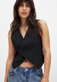 Schwarzes ärmelloses Wickeltop mit einem tiefen V-Ausschnitt und einem einzigen runden Knopfverschluss, kombiniert mit hochgeschnittenen blauen Jeans.