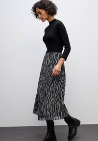 Schwarzes Langarmoberteil kombiniert mit einem grauen Rock mit schwarz-weißem Zebra-Print-Muster. Das Model trägt schwarze Stiefeletten. Schlichter Hintergrund.