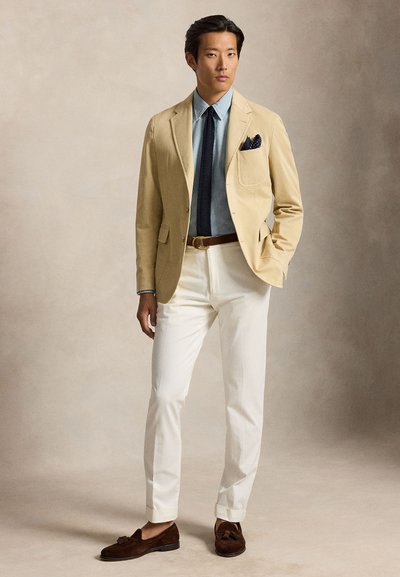 Homme portant un blazer beige, une chemise bleu clair, une cravate sombre, un pantalon blanc, une ceinture marron et des mocassins marron, debout avec une main dans la poche.