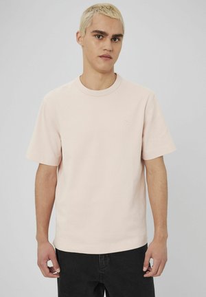 T-shirt rose clair à manches courtes en coton, avec un col rond et un logo discret sur la poitrine, porté avec un jean noir.
