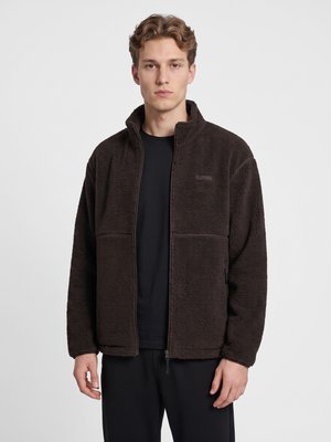 Junger Mann mit kurzen welligen Haaren, der eine schwarze Fleecejacke mit Reißverschluss und schwarze Hosen vor einem schlichten hellen Hintergrund trägt.