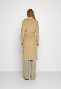 Beige langer Mantel mit taillierter Gürtellinie, eingeschnittenem Kragen und seitlichen Schlitzen, aus glattem Stoff. Über hellen, weit geschnittenen Hosen getragen.