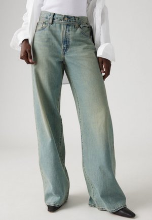 Personne portant un jean large ample bleu clair, un haut blanc et des chaussures noires pointues, debout devant un fond clair uni.