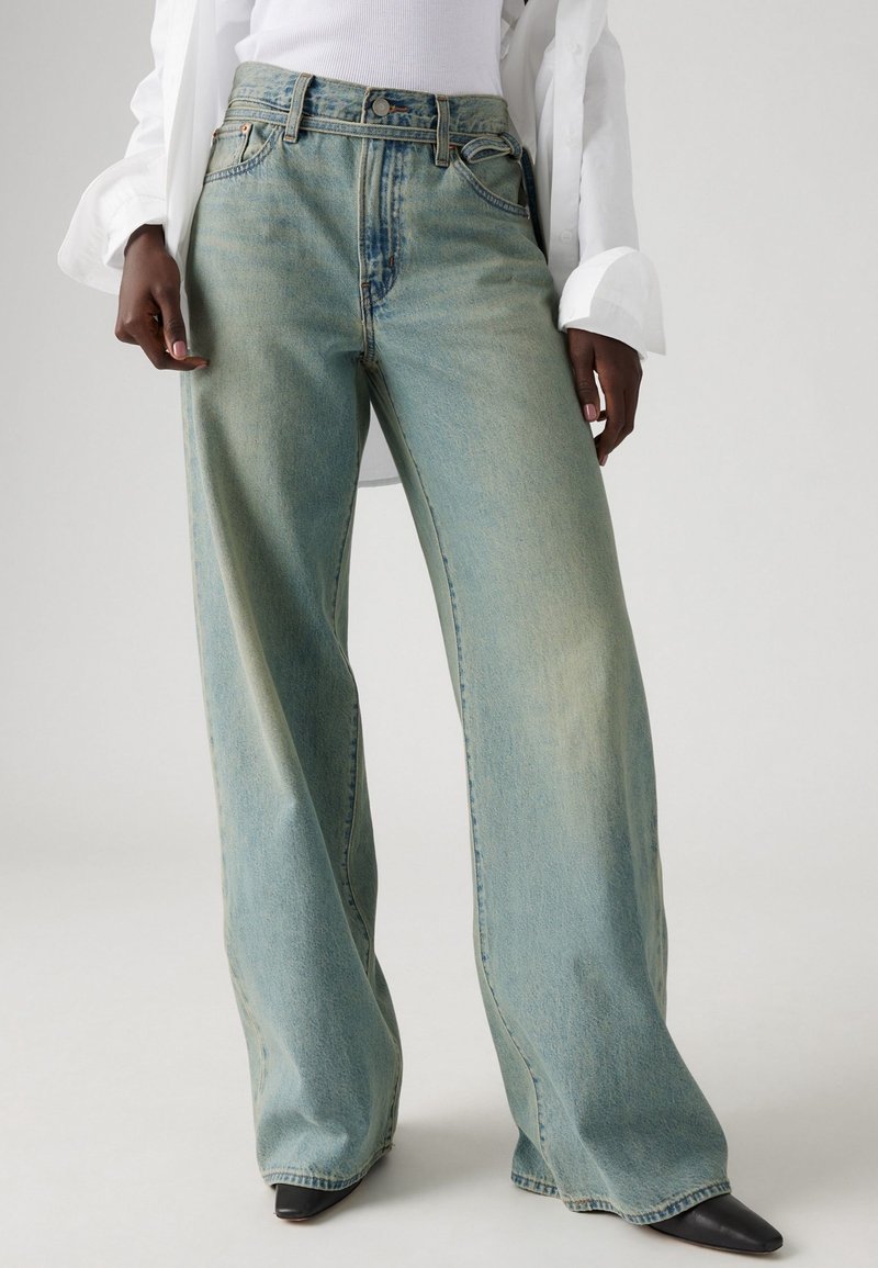Personne portant un jean large ample bleu clair, un haut blanc et des chaussures noires pointues, debout devant un fond clair uni.