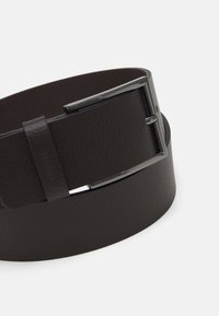 Ceinture en cuir brun avec une finition texturée, dotée d'une boucle métallique et d'une anse. Le design est élégant et contemporain.
