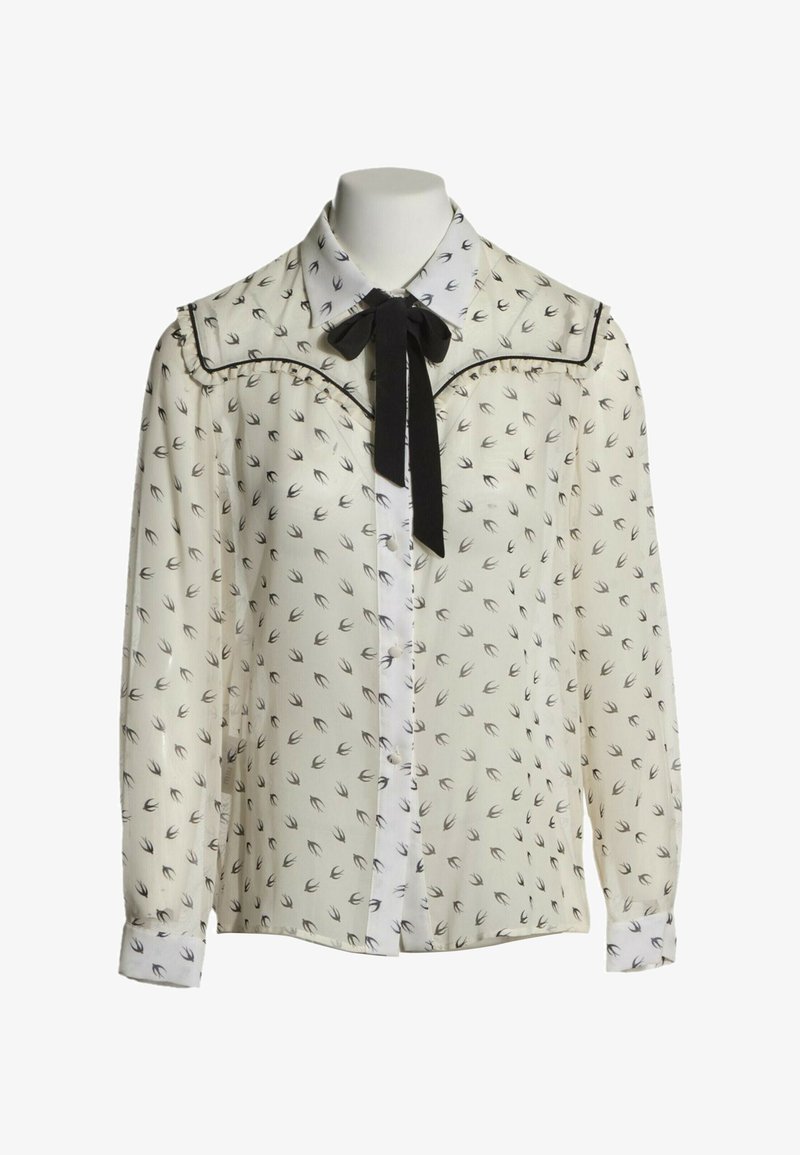 Blouse blanche à manches longues avec imprimé d'oiseaux noirs, nœud papillon noir au col et bord festonné avec un passepoil noir.