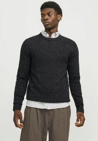 Suéter negro de punto texturizado con cuello redondo y puños acanalados, llevado sobre una camisa a rayas blanca y gris, combinado con pantalones khaki holgados.