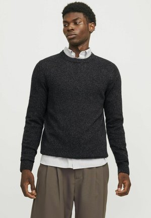 Zwarte, gestructureerde gebreide trui met een ronde hals en ribbels aan de mouwen, gedragen over een wit-met-grijze gestreepte shirt, gecombineerd met losse kaki broeken.