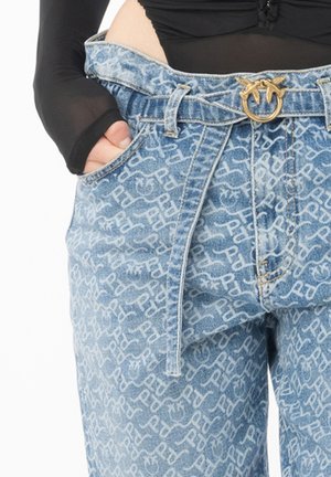 Persona que lleva vaqueros azules con un patrón, un cinturón con una hebilla dorada en forma de dos pájaros, mano en el bolsillo y una camiseta negra de manga larga.