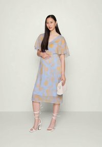 Vestido azul claro com mangas soltas, apresentando um padrão em aquarela amarelo-laranja. O modelo segura uma clutch de cor clara e usa saltos com tiras.