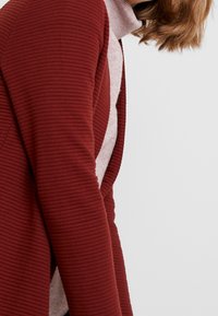 Cardigan côtelé bordeaux superposé sur un col roulé léger, présentant une texture douce et un design à devant ouvert.