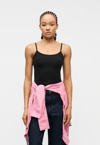 Νεαρή γυναίκα με πλεξουδωτά μαλλιά, φορώντας μαύρο αμάνικο τοπ, σκούρο τζιν και ροζ ριγέ πουκάμισο δεμένο γύρω από τη μέση της, μπροστά σε λευκό φόντο.