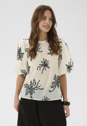 Pruunide pikkade juustega naine, kes kannab valget pluusi musta lillemustrilise tikandi ja padjandunud varrukatega, mustade pükste ja musta käevõruga.