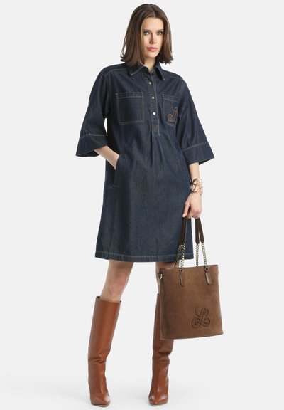 Denim-paitamekko, jossa on lyhyet hihat, kaksi rintataskua ja ruskeat ompeleet. Yhdistettynä pitkiin ruskeisiin saappaisiin ja ruskeaan nahkalaukkuun.
