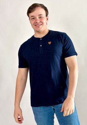 Jeune homme souriant, portant un t-shirt henley bleu marine à manches courtes et un jean bleu, debout devant un fond clair uni.