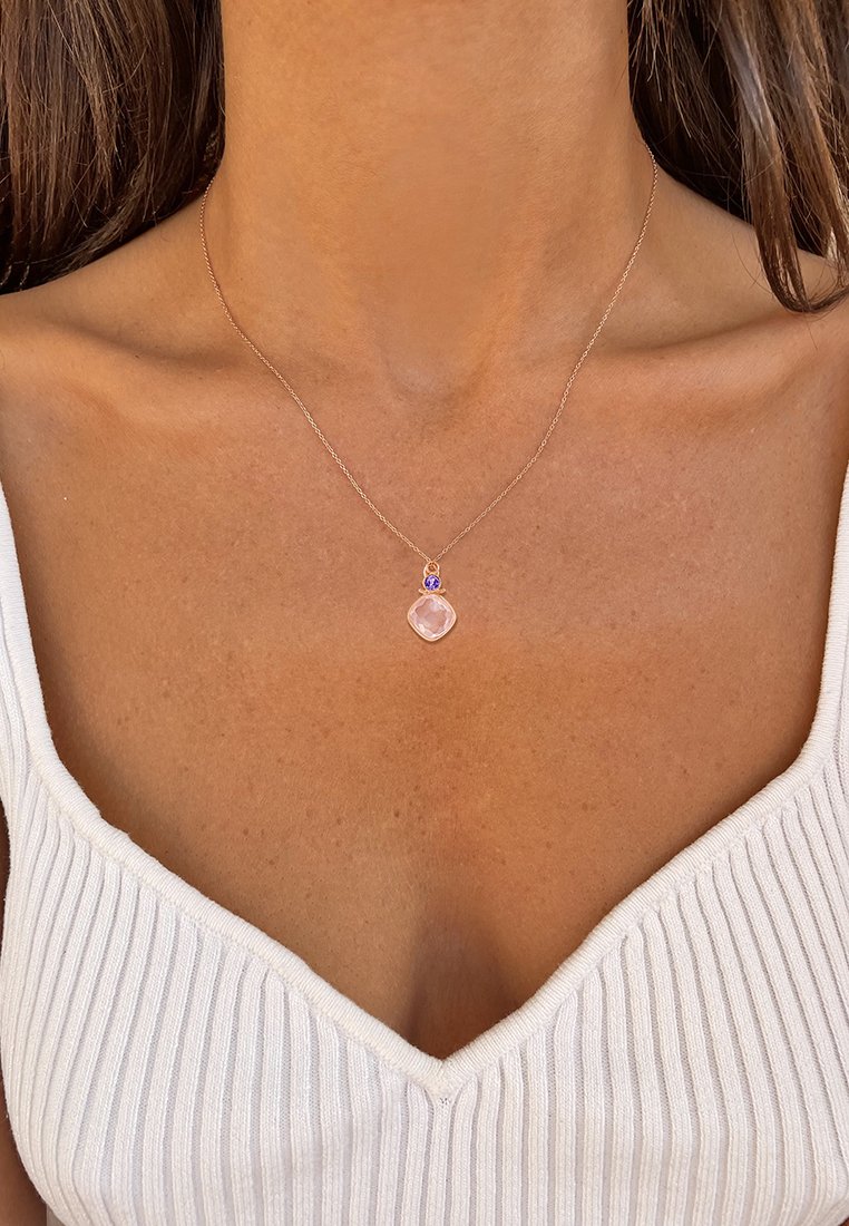 Collana in oro rosa con un pendente trasparente e sfaccettato, arricchito da una pietra di accentuazione viola, sospeso a una catena sottile. Sfondo texturizzato nella parte superiore.