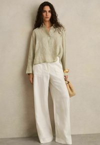 Leinenhemd in Hellgrün, knöpfbar mit Kragen, kombiniert mit weiten weißen Leinenhosen. Die Accessoires umfassen eine beige geflochtene Tasche.