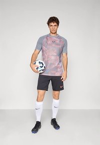 Camisa deportiva gris y roja con estampado, pantalones cortos negros, calcetines blancos y botas de fútbol, sosteniendo un balón de fútbol. Logotipo notable en la camiseta.