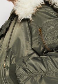 Chaqueta bomber verde oliva con un suave collar de piel sintética blanca, que presenta un bolsillo con cremallera y una tela texturizada con un acabado liso.
