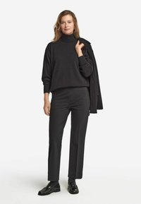 Svart polotröja med ribbad ärm, tillsammans med skräddarsydda svarta byxor och glänsande svarta skor. Modellen håller en svart jacka.