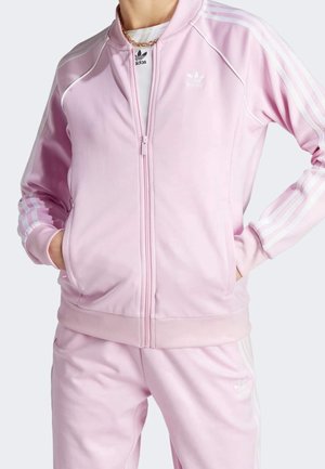 Osoba ubrana w jasnoróżowy dres Adidas z białymi paskami i logo, ręce w kieszeniach, pod spodem widoczna biała bluzka, złoty łańcuszek na szyi.