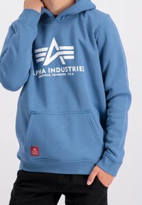 Felpa blu con cappuccio e tasca frontale, caratterizzata da un logo e testo bianchi. Realizzata in tessuto morbido con polsini e orlo a coste. Etichetta rossa sul lato.