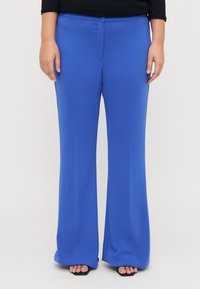 Pantalons larges bleus en tissu lisse et léger avec un fermoir à l'avant et des détails de plis subtils. Chaussures noires visibles en dessous.