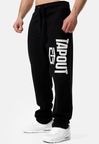Fekete, puha anyagból készült melegítőnadrág széles derékkal, zsinórral, oldalzsebekkel és jól látható fehér „TAPOUT” logóval.
