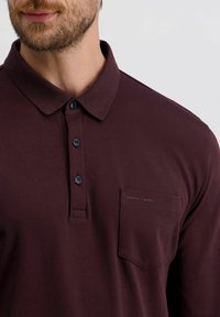 Mann trägt ein dunkelburgunderfarbenes Poloshirt mit drei Knöpfen und einer Brusttasche, die mit "pierre cardin" beschriftet ist.
